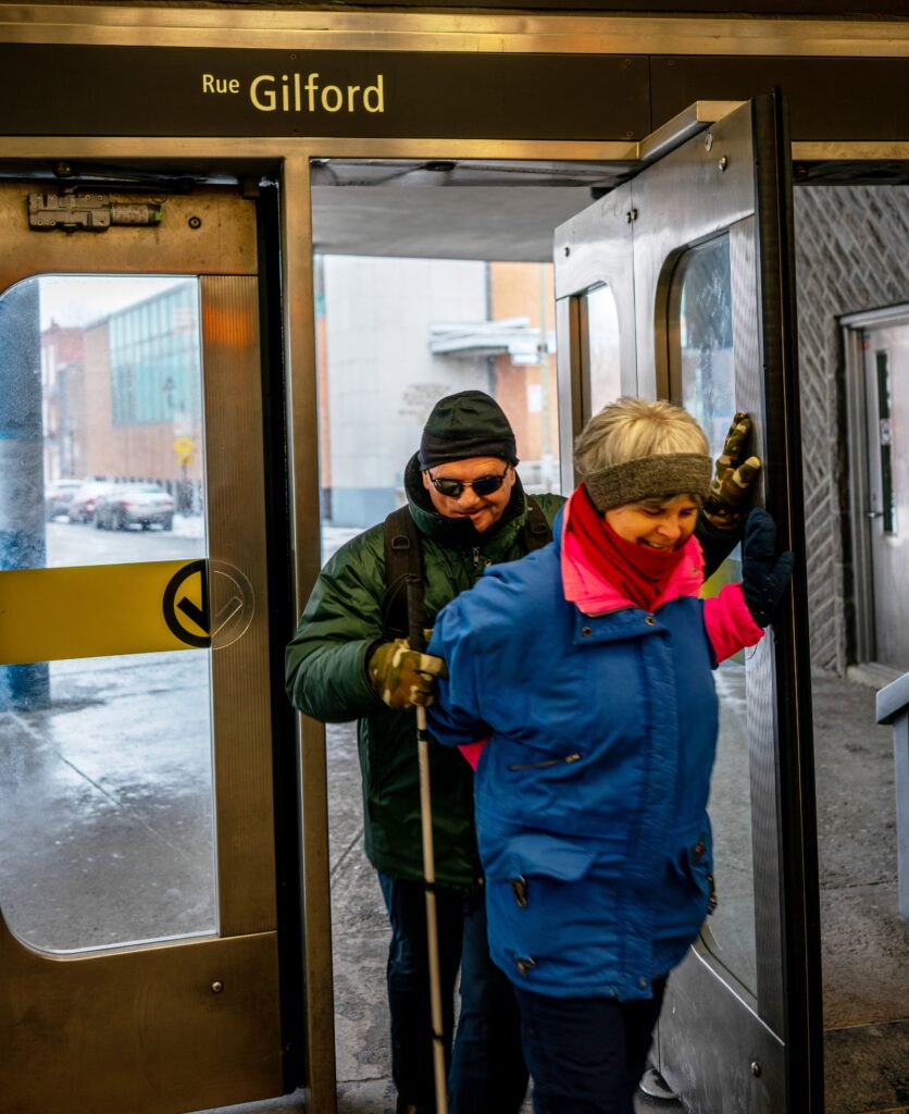 Service d'aide bénévole : Un homme aveugle est accompagné par une bénévole dans le métro.